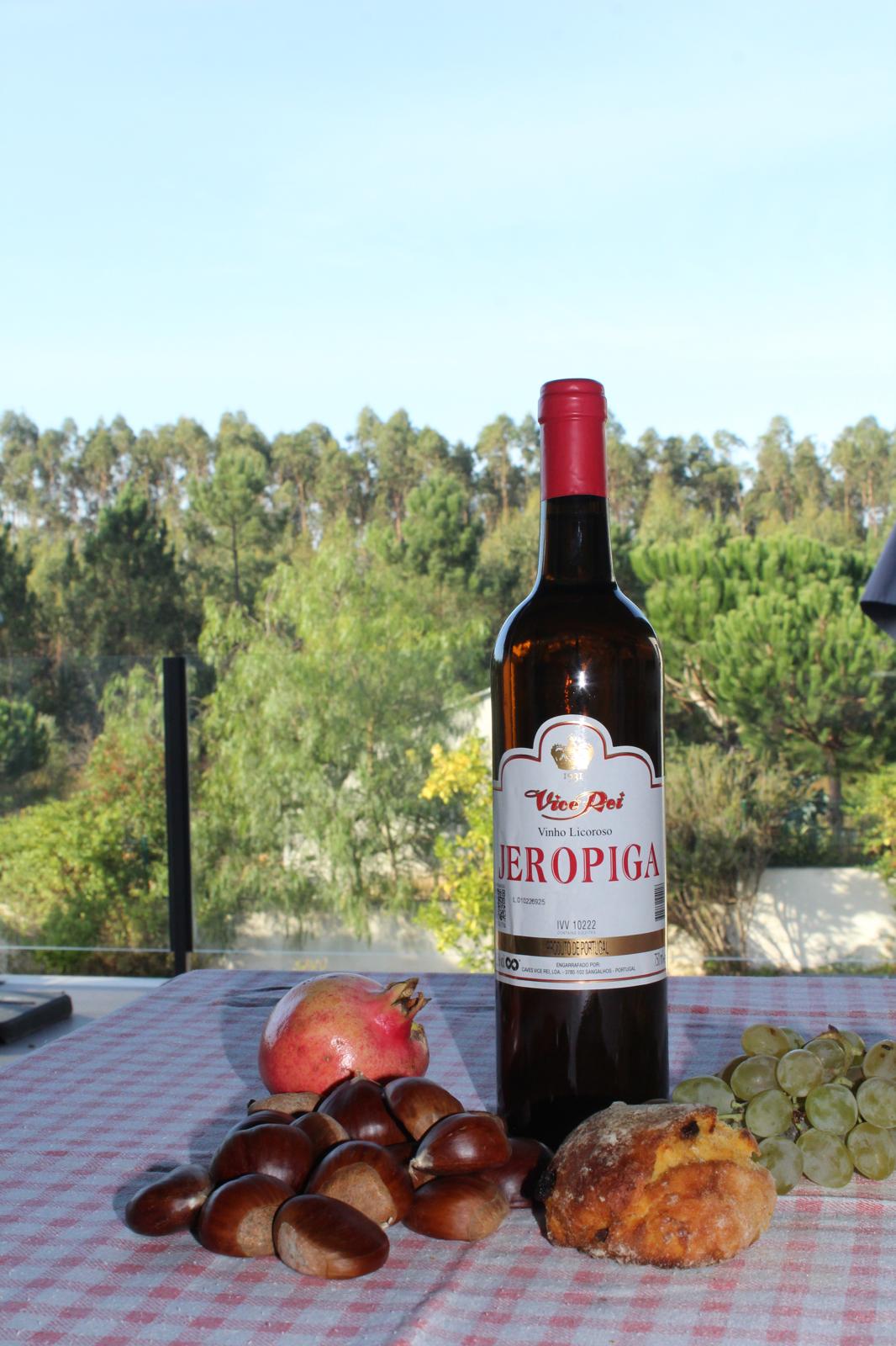 Bottle of JEROPIGA TRADICIONAL VICE REI 0.75 surrounded by chestnuts, pomegranate, and grapes on a checkered tablecloth.