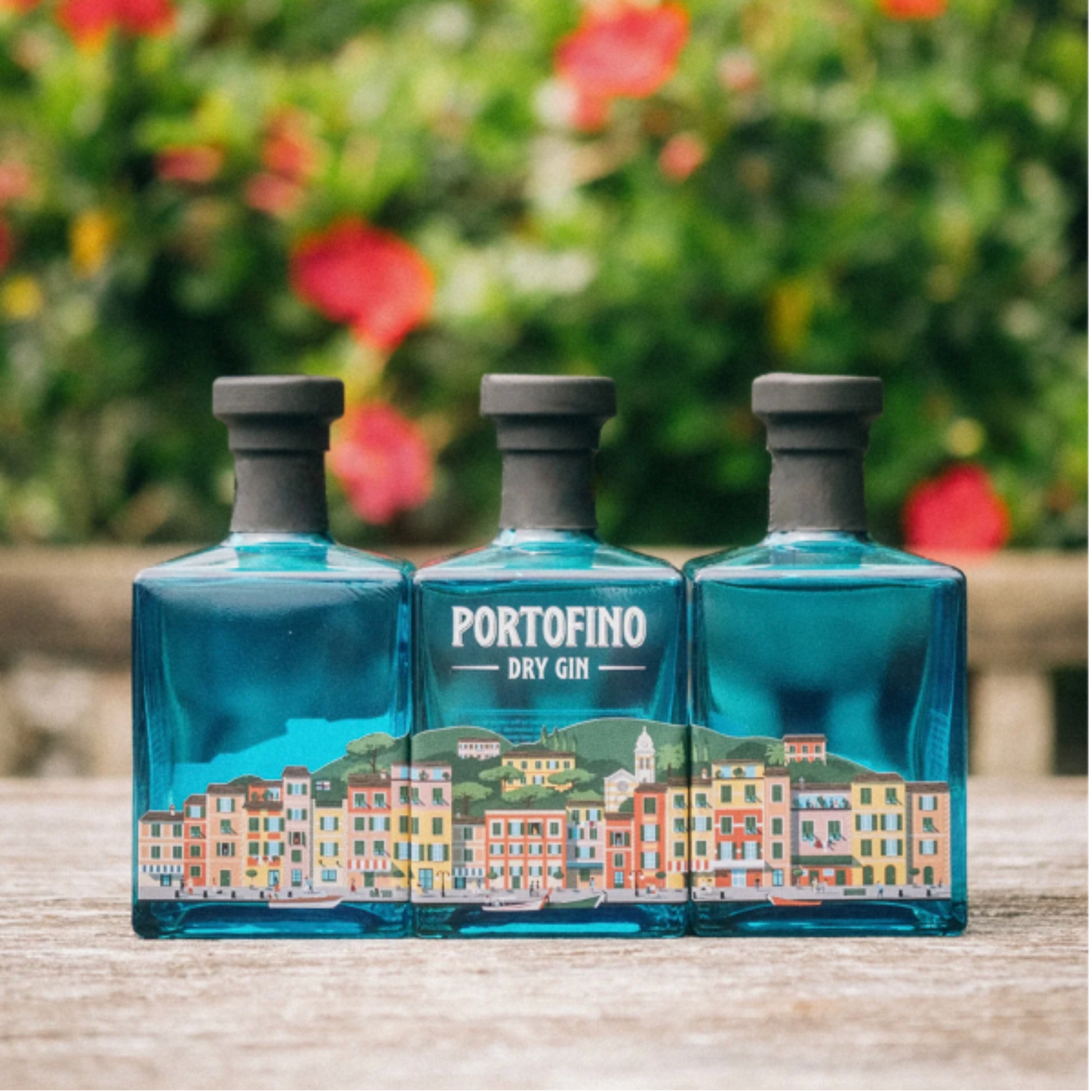 Três garrafas de PORTOFINO DRY GIN DE ITÁLIA 0.50L com rótulo colorido, em frente a um fundo florido.
