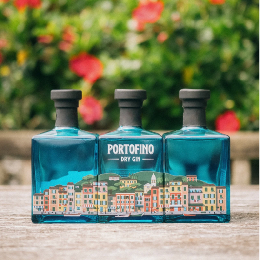 Três garrafas de PORTOFINO DRY GIN DE ITÁLIA 0.50L com rótulo colorido, em frente a um fundo florido.