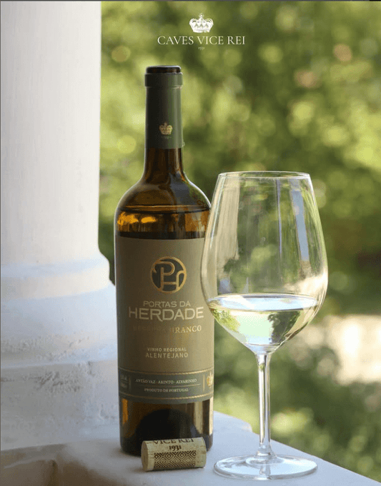 Bottle of Portas da Herdade Branco Reserva 2024 with a glass of white wine, set against a blurred green background.